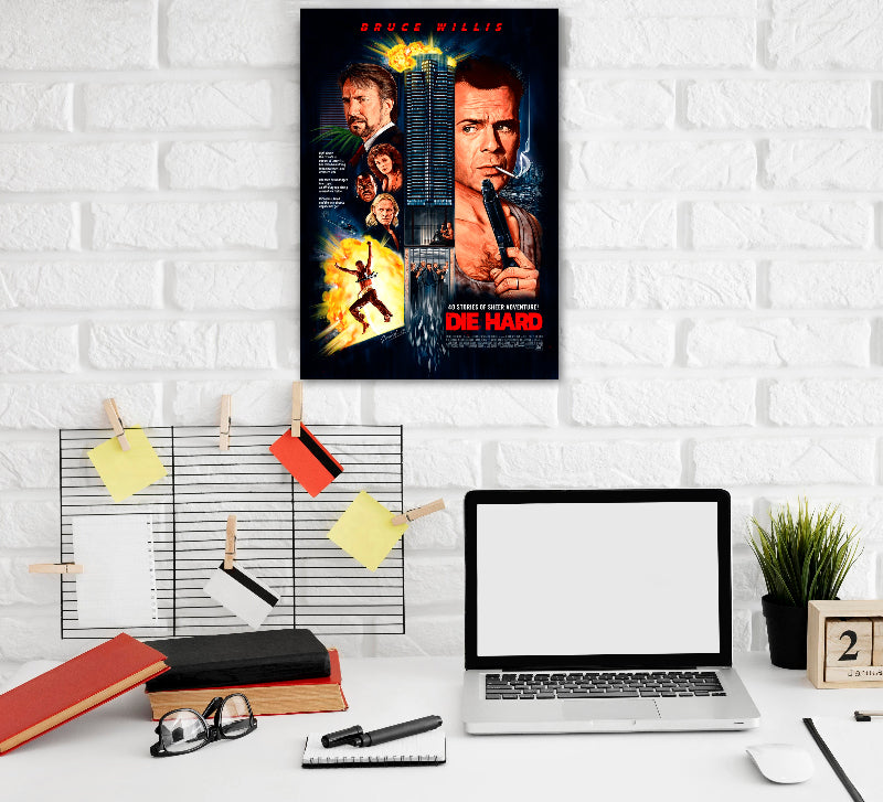 A "Die Hard" movie poster featuring Bruce Willis is mounted on a white brick wall above a minimalist workspace. The poster shows Bruce Willis holding a gun, scenes of explosions, and other characters from the film, with the Nakatomi Plaza skyscraper in the center. Below the poster, the workspace has a laptop, books, glasses, sticky notes clipped on a metal grid, a plant, Action Thriller Hollywood Movies Adventure