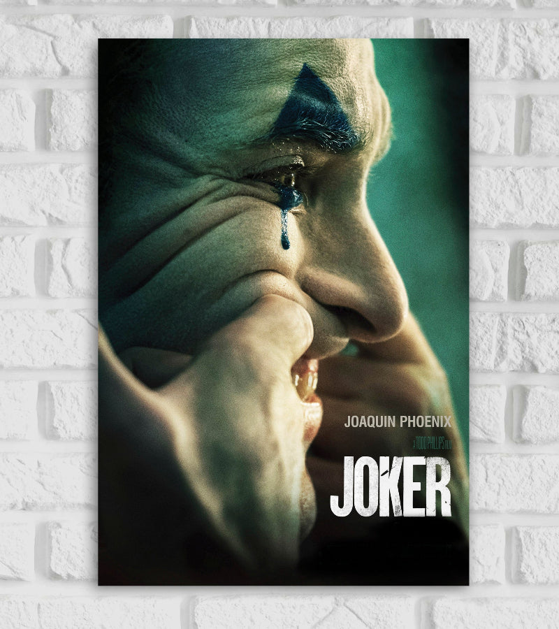 Posters Frame Of Hollywood Action Drama Thriller Crime Arthur Fleck The close-up side profile shows the Joker forcing a smile with his fingers while a single tear streaks down from his eye, smudging his clown makeup. His expression is somber and haunting. The background is muted green, creating a moody and dramatic atmosphere. The title "JOKER" appears in bold white text at the bottom right, with "Joaquin Phoenix" and "A Todd Phillips Film" above it.