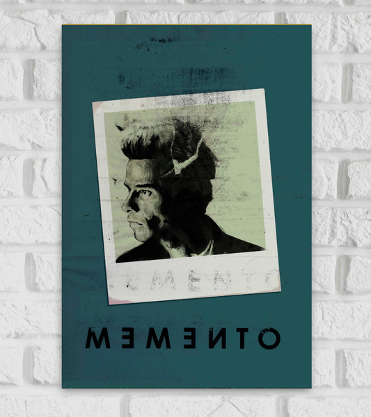 Poster for the psychological thriller "Memento." The design features a distressed, faded Polaroid photo tilted slightly, showing a black-and-white portrait of a man looking to the left with a serious expression. The background is a dark teal color, and the title "MEMENTO" is written at the bottom in black, but flipped horizontally, creating a mirrored effect. The worn, textured look of the image emphasizes the film's themes of memory loss, confusion, and fragmented reality American Hollywood Action