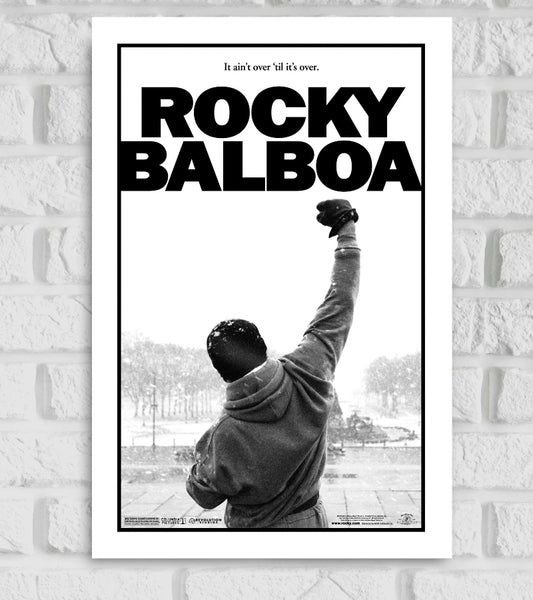Black and white poster of the movie 'Rocky Balboa' featuring the iconic image of Rocky, wearing a hoodie, with his back to the viewer and one fist raised triumphantly in the air. The background shows a snowy cityscape, and the text at the top reads 'It ain't over 'til it's over.' Bold black text in the center says 'ROCKY BALBOA'