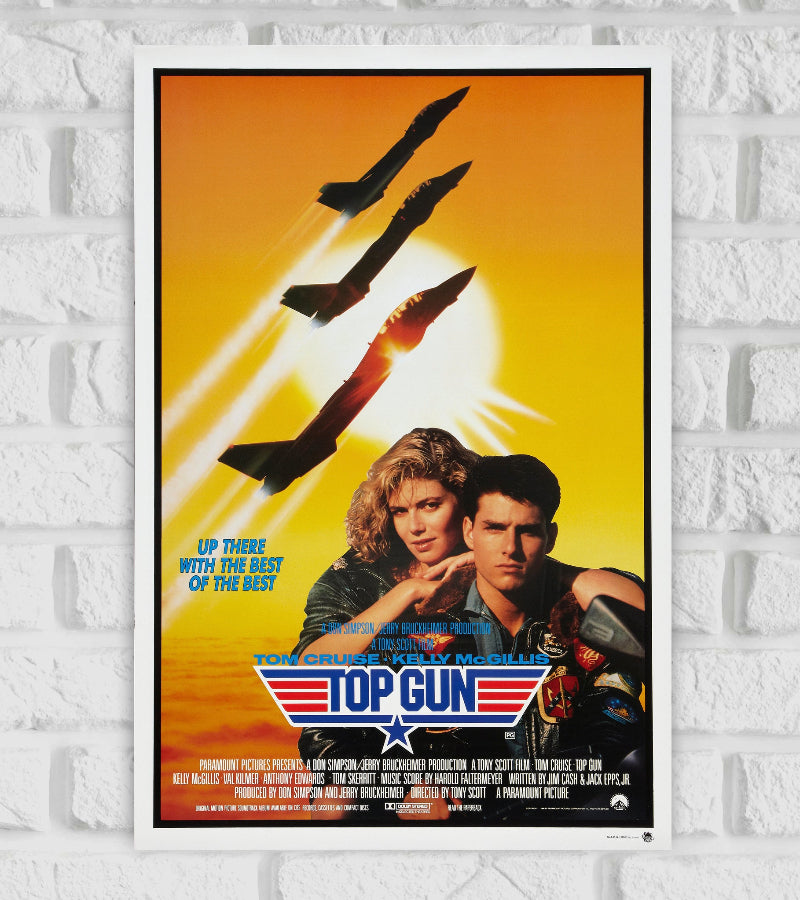 Poster of Top Gun featuring Tom Cruise and Kelly McGillis in the foreground, with Cruise wearing a flight jacket and intense expression. Behind them, three fighter jets soar upward into the sky, leaving white contrails against a bright yellow sunset. The film’s iconic logo with wings and a star is placed below them, along with the tagline "Up there with the best of the best." The design evokes themes of aviation, action, and romance Hollywood Actor Actress Action Drama Romance