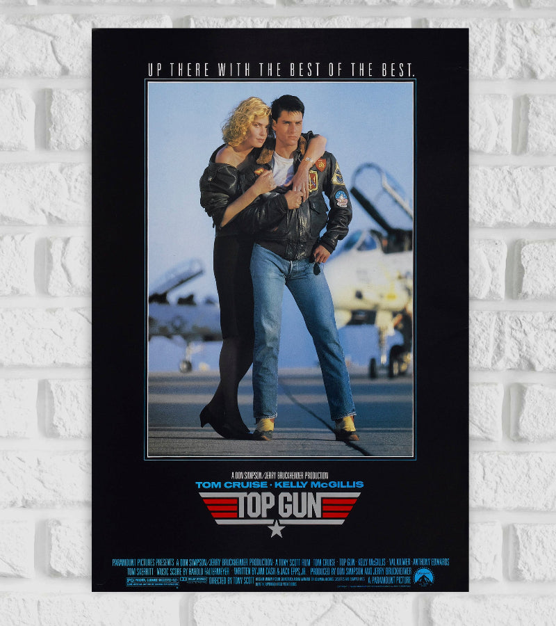  Poster of the movie "Top Gun" featuring Tom Cruise and Kelly McGillis standing closely together on an airbase, with fighter jets visible in the background. Tom Cruise wears a leather pilot jacket and jeans, while Kelly McGillis wears a black outfit and rests her hand on his shoulder. The top of the poster reads "Up there with the best of the best." The title "Top Gun" with a red and silver emblem Hollywood Adventure Action Drama Romance Actress Actor 