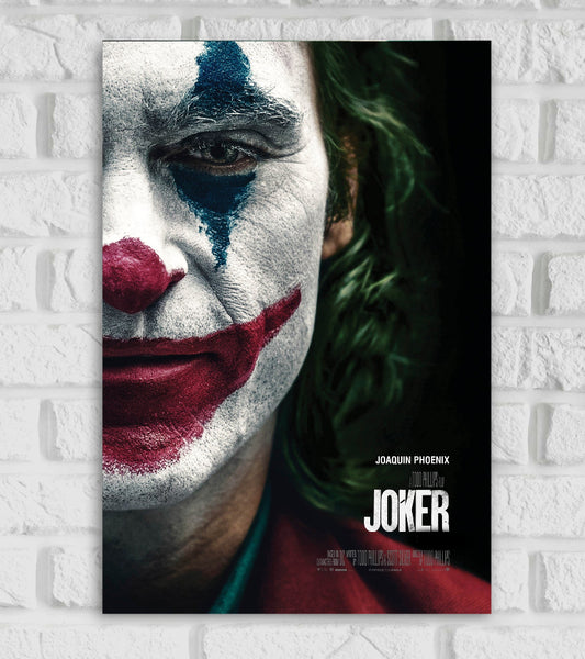 Poster of the movie 'Joker' featuring a close-up of Joaquin Phoenix's face in full clown makeup. His expression is somber, with smeared red lipstick forming a forced smile and blue paint above and below one eye. The background is dark, emphasizing the character’s green hair and red suit. The title 'JOKER' is displayed in bold white text at the bottom right, with Joaquin Phoenix’s name above it. The poster is mounted on a white brick wall."









